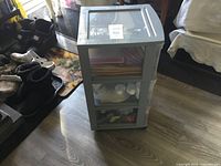 Three plastic storage drawer units standing upright, containing craft supplies such as paper, glue bottles, paint, and brushes inside translucent drawers.