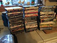 Three stacks of DVD movies in original cases displayed on a table by a window. Variety of titles visible on cases.