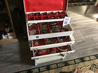 Top-down view of white three-tier jewelry box with red interior displaying multiple compartments filled with vintage jewelry pieces including earrings, bracelets, and rings.