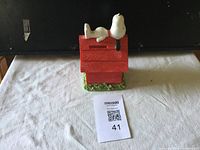 Front view of the Snoopy ceramic bank showing Snoopy lying on top of the red doghouse with green base.