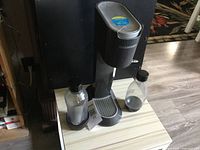 SodaStream soda maker and two clear plastic bottles on a white table with dark background showing the machine and bottles from front angle.