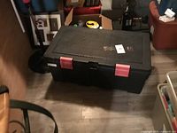 Closed large black plastic tool box with red latches on a wooden floor surrounded by other household items.