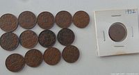 Photo of 16 Canadian pennies, mostly from the 1920s and early 1930s, arranged on a surface including one coin encased in a white cardboard holder labeled '1932'.