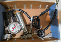 Grease gun device showing metal handle, pressure gauges, and hoses inside a cardboard box
