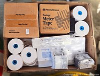 Top view of a box containing multiple rolls of paper with blue cores, box of Pitney Bowes Postage Meter Tape, and various sealed packets.