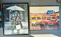 Both wall art pieces side by side. The left piece shows the black and white photo of two girls under an umbrella. The right piece shows the colorful malt shop scene with classic cars. The malt shop frame has visible damage.