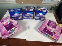 Photo showing five packages of bladder protection pads: three unopened Tena Overnight packages in purple and blue, and two pink Poise pad packages, one partially open.