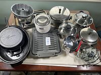 Overview of all kitchenware items arranged on table including pots, pans, lids, enamel roaster, rice cooker, broiling pan, baking molds.