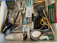 Top-down view of drawer full of assorted kitchen utensils including peelers, whisks, ladles, tongs, scissors, brushes, and ceramic spoon rest.