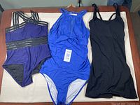 Three large bathing suits laid out on a table: dark blue with black sheer stripes, royal blue halter neck, and simple black one-piece.