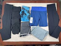Six new women's bottoms in medium size displayed on a flat surface. Items include leggings, biker shorts, and shorts in black, blue patterned, turquoise, and light grey colors.