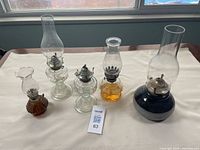 Assortment of five oil lamps displayed on a white surface against window light, showing height and detail variety.