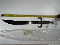 Full view of the silver scimitar next to a measuring tape showing length of about 21 inches along with its plastic protective covering.