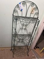 Full view of the black metal wire rack with three wire shelves and decorative scrollwork against a wall.