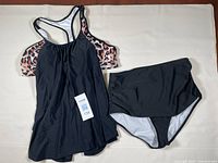 Photo shows a two-piece women's bathing suit laid flat on a light surface. The top is a black tankini with leopard print on the straps and sides and a racerback design. The bottom is a high-waisted solid black bathing suit bottom with white lining. A size tag visible reads XL.
