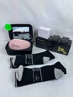 Pink Eyeology handheld massager resting inside a black zippered case, with a charger cable coiled inside the case. On right side of image, pair of black and grey Dr. Warm heated socks laid flat in front of a black and gold Dr. Warm branded box along with two small black boxes and one small plain white box, presumably accessories or chargers for the heated socks.