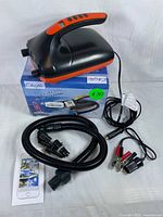 Photo showing black and orange Chefic electric air pump on top of its original box. The box shows branding and indicates 12V power. Accessories visible include flexible hose, multiple nozzle adapters, power cable with alligator clips.