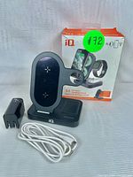 Charging stand positioned with power adapter, USB cable, and original packaging box with iQ branding visible. The stand shows designated wireless charging areas for phone, smartwatch, and earbuds.