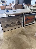 Two framed art prints on floor beneath a folding table. One is a black and white photo of a couple on a beach, the other is a colorful scenic print with a lighthouse at sunset.