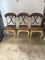 Three wooden dining chairs aligned side by side showing full front view. Each chair features dark stained seat and back with cream painted cabriole legs. Shows wear from use.