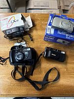 Four cameras on wooden surface: Canon Snappy Alpha IX II in original box, Olympus D-380 digital camera in box, Vivitar Series 1 camera, and Vivitar PS30 camera.