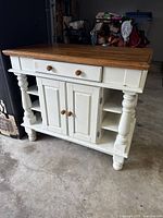 Front angled view showing entire wooden island with white base, turned legs, double cupboard doors and drawer with wooden knobs.