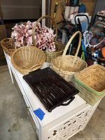 Photo showing assorted wicker and woven baskets in natural material alongside a pink artificial flower wreath, and a small dark wooden tray with handles.