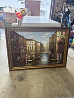 Front view of framed oil painting showing Venice canal scene with gondolas and architectural details, set on the floor with visible background items