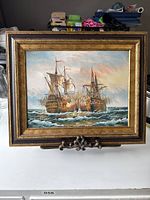 Front view of the framed oil painting showing two ships on rough seas with sky background, placed on ornate metal stand.