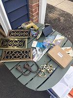 Wide view of hardware and tools on a green table outside, including brass vents, power drill, curtain rods, hooks, small hardware, and a doorbell.