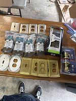 Several packs of Liberty brand bronze cabinet knobs (10 per pack), assorted brass and brushed nickel wall plates and outlet covers arranged on table, including decorative plates, and new packaged Schlage door lockset with brass knobs.