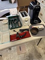 Full view of kitchen items on a table including white toaster, Nespresso coffee machine, cutlery set in green case, glass bake dishes, small saucepan, espresso maker, and red tray