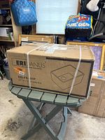 Photo shows closed cardboard box with ERIDANUS branding and sketch of utility sink, on green folding table in storage area.