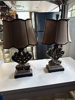 Pair of table lamps approximately 24 inches tall with ornate carved bases and brown fabric shades taken on a white table with kitchen background