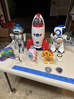 Full layout of toys including silver robot, white and red rocket with astronauts, white and blue robot, pink elephant, yellow lion figure, remote and accessories on table.