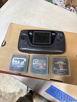 Top view of Sega Game Gear with 3 game cartridges in clear cases (Columns, Super Monaco GP, Clutch Hitter) on a cardboard surface.