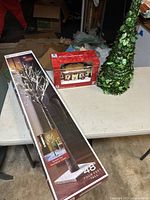 Wide angle showing all three Christmas decor items on a white table including boxed lighted tree, boxed village building, and leafy tree