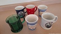 Front view of all six mugs arranged on counter, showcasing color and logo variety including green glass mug and Oilers logos.