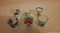 Six assorted souvenir shot glasses displayed on a countertop, showing different city and travel-themed graphics including Boston and cartoon lobster figure