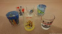 Six assorted souvenir shot glasses arranged on a counter, showing various city-themed colorful designs including New Orleans and Montreal.