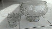 Clear glass punch bowl with grape pattern next to six small matching cups on tiled floor