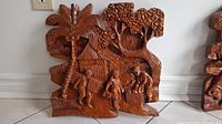 Photo of a medium-sized wood relief carving showing a village scene with three people, a hut, and trees.