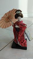 Front left angle view of Japanese Geisha doll holding peach-colored umbrella, wearing red floral kimono and blue obi sash, mounted on black base.