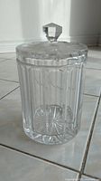 Full view of the medium clear glass container standing upright on tiled floor, showing vertical and oval cut glass patterns around body.