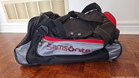 Side view of the Samsonite duffel bag showing fabric material, black, red, and gray colors, and Samsonite logo on the side pocket
