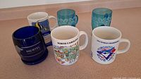 Set of 6 assorted mugs including glass and ceramic types on a countertop showing Nortel, North Carolina, Toronto Blue Jays logos and blue glass mugs with snowflake patterns.