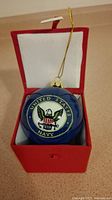Blue round glass Christmas ornament with United States Navy emblem inside a red box with white lining, gold hanging string visible.