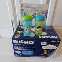 Two President's Choice insulated spill-resistant cups with green and blue lids, placed on top of a sealed Huggies Overnites diapers case.