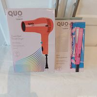 Photo of the boxed Quo by Conair orange travel hair dryer and pink mini straightener in their retail packaging displayed on a white surface.