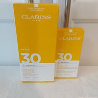 Two boxes of Clarins sunscreens, one larger body cream box and one smaller facial sunscreen box, both with SPF 30 Broad Spectrum protection, yellow and white packaging.